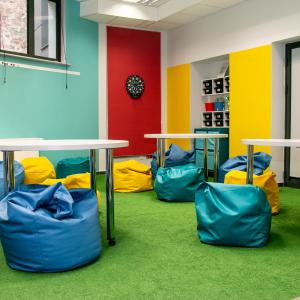 Widok wnetrza sali Roszarnia ze ścianami w kolorze żółtym, turkusowym i malinowym oraz wykładziną ala trawa. Widok na okrągłe stoły i kolorowe pufy do siedzenia.