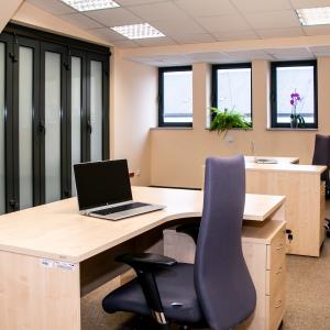 Widok wnętrza pomieszczenia biurowego z ujęciem spod ściany z za jednego z biurek, skosem w kierunku otwieranej ściany oraz okna. Na zdjęciu dwa biurka oraz fotele obrotowe, na parapetach kwiaty.